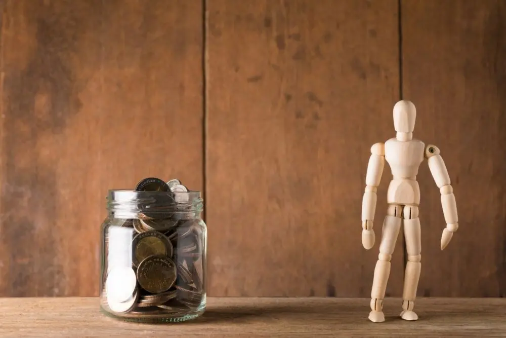 Jar of coins next to a wooden mannequin, showing money set aside for an emergency fund