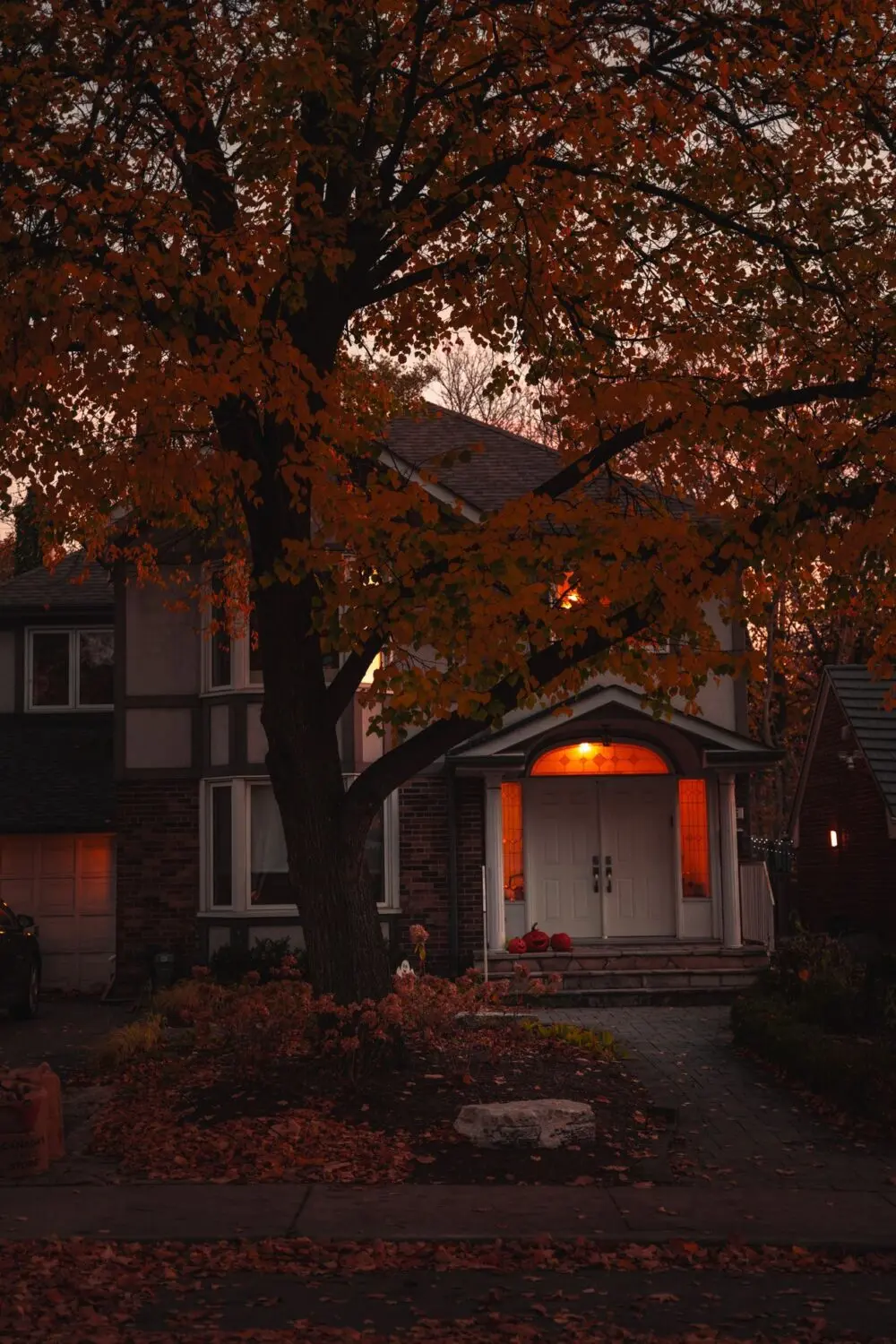 House with a lit porch at dusk under autumn trees, as early sunsets arrive