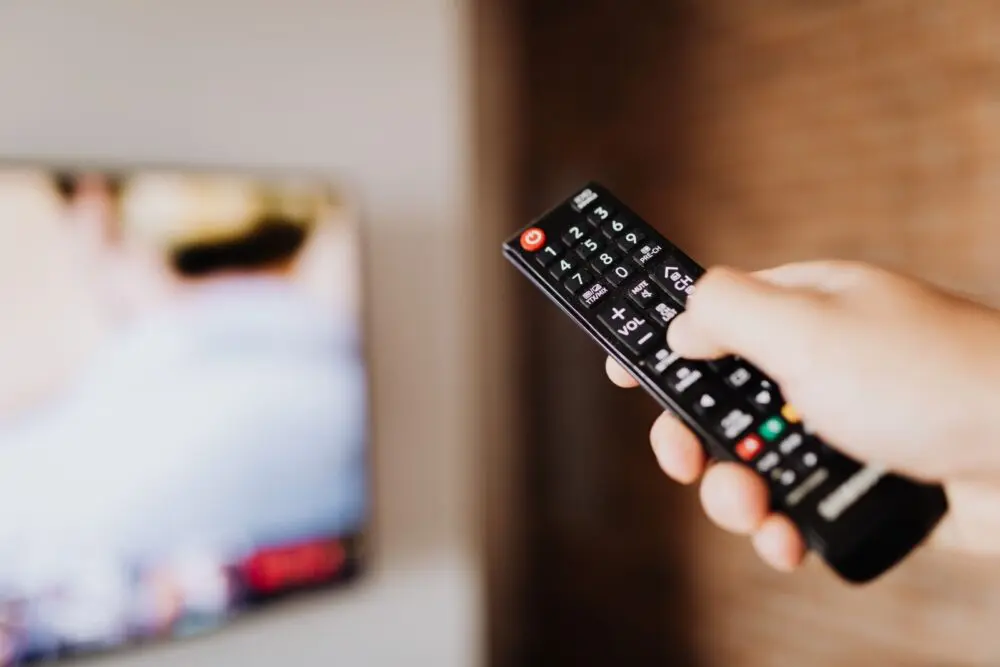 Hand holding a remote control in front of a TV, ready to turn off smart TV tracking in settings