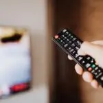 Hand holding a remote control in front of a TV, ready to turn off smart TV tracking in settings