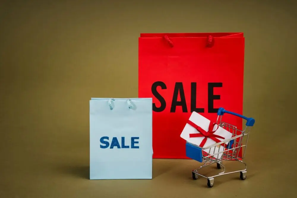 Sale shopping bags and a mini cart holding a wrapped gift, for holiday shoppers on a budget