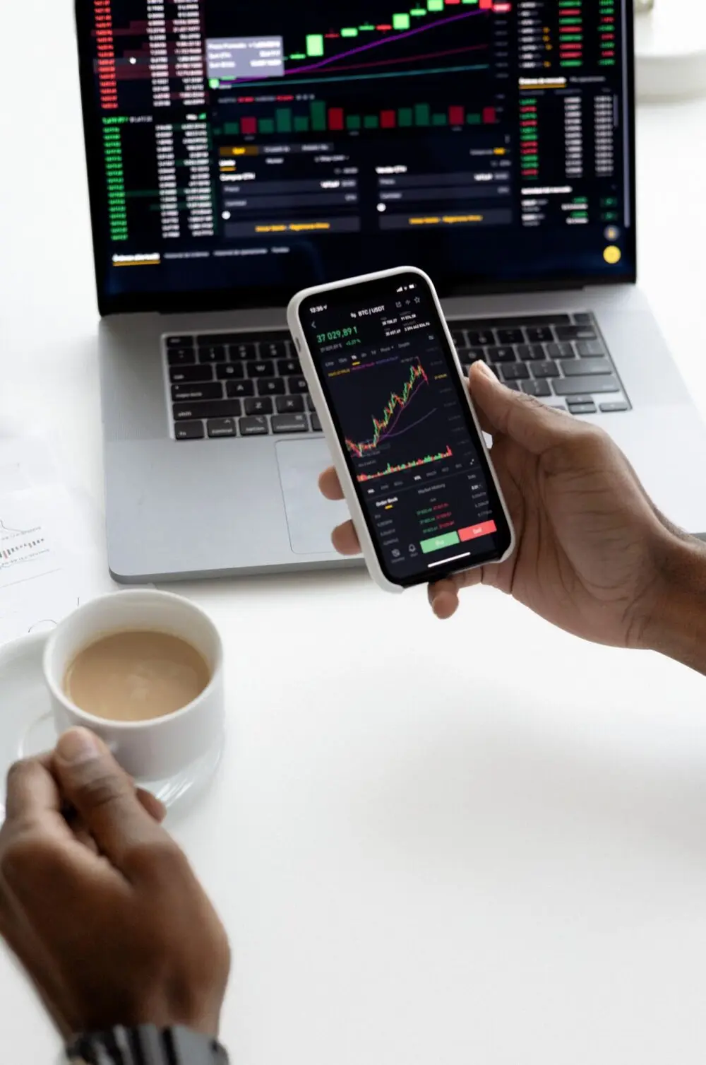A hand holding a phone that shows a financial chart beside a laptop and coffee
