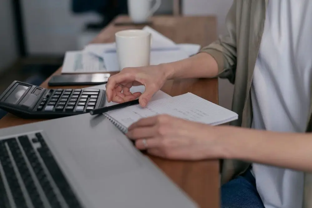 Hand writing in a notebook beside a calculator while preparing to create an invoice