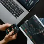Hands holding a phone showing a market chart beside a laptop and printed charts