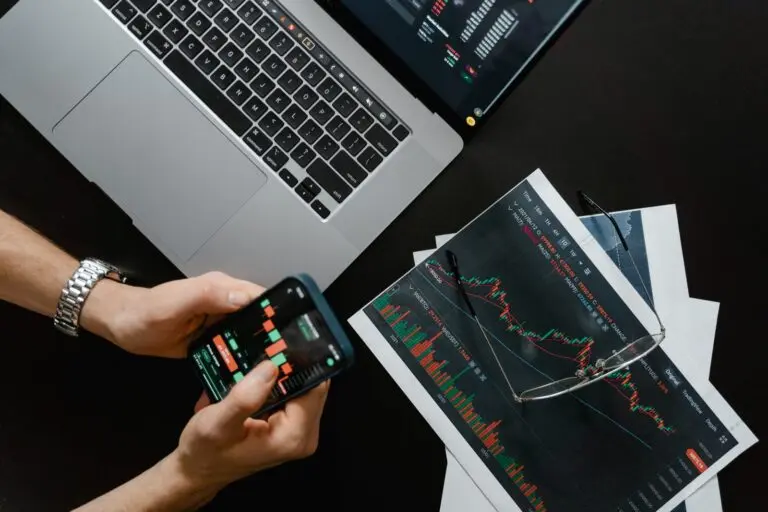 Hands holding a phone showing a market chart beside a laptop and printed charts