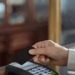 Hand holding a black credit card over a payment terminal, a Capital One earnings theme